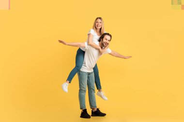 Neşeli bin yıllık beyaz bir adam beyaz tişörtlü kadını sırtında sürüklüyor, anın tadını çıkarıyor, sarı arka planda izole edilmiş, tam boy. Çiftler boş vakitlerinde eğlenirler, reklam ve teklif, ilişki ve aşk.