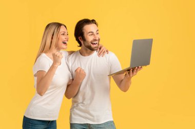 Beyaz tişörtlü neşeli beyaz beyaz bir çift ellerinde dizüstü bilgisayarlarıyla, sarı arka planda izole edilmiş zafer işareti yapıyorlar. Başarılı bir anlaşma, online kazanç, harika bir teklif ve satış, stüdyo
