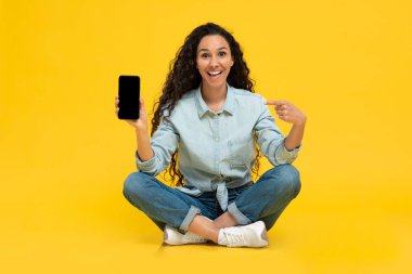Neşeli genç bayan tasarım için akıllı telefonu işaret ediyor, sarı stüdyo arka planında yeni uygulama veya web sitesi tanıtıyor, tam uzunlukta. Cep telefonu görüntüleme şablonu, ilanınız için boşluk