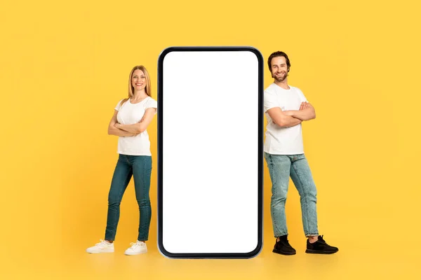 Excellent digital ad and offer. Cheerful millennial caucasian couple in white t-shirts standing near huge phone with empty screen, isolated on yellow background, full length. New app, website and blog
