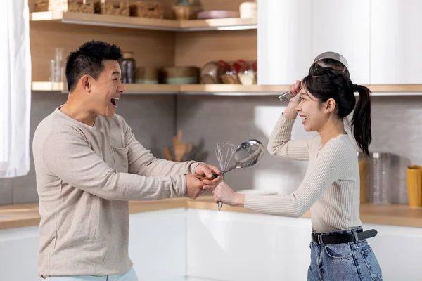 Funny japanese couple having good time while cooking together at home, positive asian man and woman in casual outfits holding kitchen utensils and making fight, imitating knights
