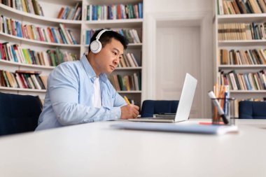 Orta yaşlı Asyalı iş adamı dizüstü bilgisayarda çalışıyor kulaklık takıyor ve ofis odasında kitap raflarıyla notlar alıyor. E-Öğrenme ve Teknoloji. Yan Görünüm