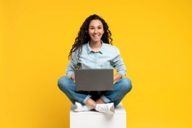 Dizüstü bilgisayarla bacak bacak üstüne atmış, kameraya gülümseyen, sarı stüdyo geçmişi üzerinde çalışan ya da ders çalışan neşeli genç bir kadın. Uzaktan eğitim, iş, iletişim konsepti