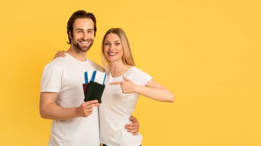 Beyaz tişörtlü neşeli beyaz adam ve kadın, sarı arka planda izole edilmiş uçak biletleriyle pasaportları işaret ediyor. Birlikte yolculuk, seyahat, covid-19 'dan sonra tatil.