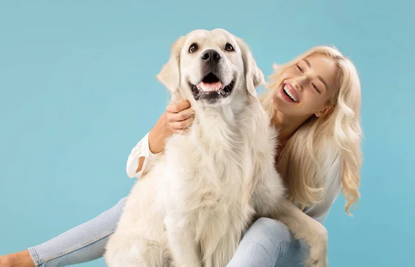 Pets and people concept. Happy young lady posing with her adorable golden retriever dog, sitting over blue studio background, embracing and looking at her pet