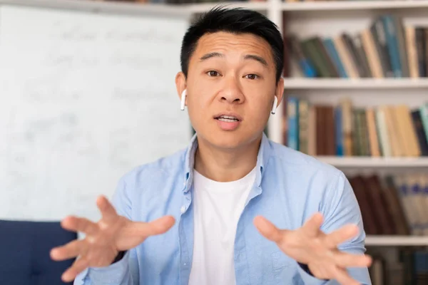 Middle Aged Korean Teacher Male Video Calling Talking To Camera Wearing Wireless Earbuds Communicating And Teaching Online Sitting In Modern Library. Internet Education Concept