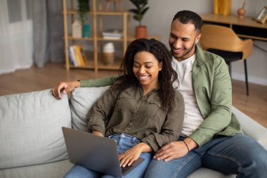 İnternette dizüstü bilgisayar kullanan mutlu Afrikalı Amerikalı çift ve hafta sonları evdeki koltukta oturup rahatlarken web sörfü yapıyorlar. Teknoloji ve Gadget Kavramı