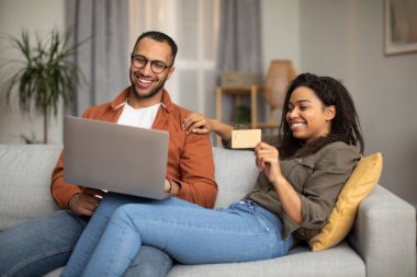 Neşeli siyah çift online alışveriş yapıyor dizüstü bilgisayar ve kredi kartı kullanarak ödemeyi evdeki koltukta yapıyor. Eşler bir şey satın alıyor. İnternet Bankacılık ve Ticaret Konsepti