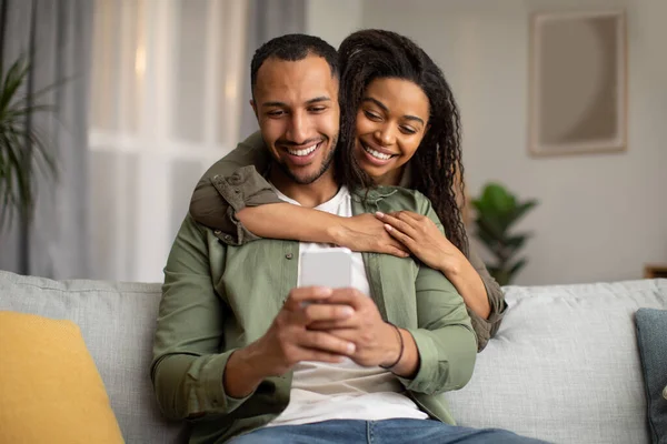 快乐的非洲裔美国夫妇使用手机浏览互联网和发短信在一起 妻子拥抱丈夫坐在沙发上在家里 配偶在手机上使用应用程序 技术概念 — 图库照片