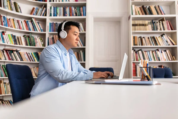 Side View Asian Man Wpisując Laptopie Noszenie Słuchawek Siedzi Bibliotece — Zdjęcie stockowe