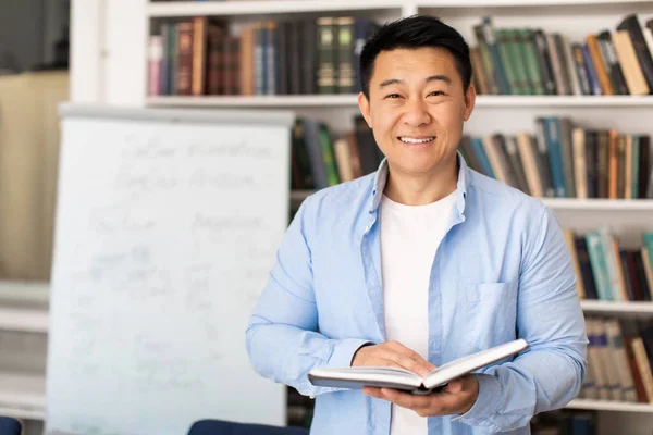 Korean Male Teacher Holding Book Standing Near Whiteboard Smililng Looking At Camera Teaching And Posing In Classroom Or Library. Knowledge And Modern Education Concept