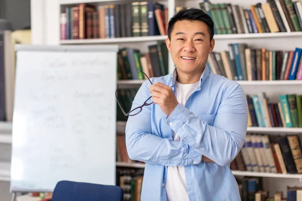 Happy Asian Male Teacher Posing Holding Eyewear Smiling To Camera Standing Near Whiteboard With English Grammar Rules And Bookshelf In Classroom Indoor. Modern Education
