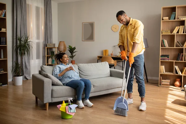 妊娠中 アフリカ系アメリカ人の夫が自宅でソファに座って携帯電話を休憩し 使用している間に家を掃除します 家族のライフスタイルと家事レジャーコンセプト — ストック写真