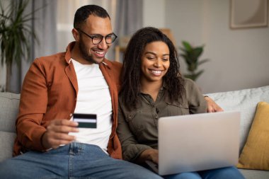 Mutlu Afro-Amerikan çift online alışveriş yapıyor dizüstü bilgisayar ve kredi kartı evde koltukta oturuyor. Bilgisayarda internet bankacılığı hizmetini kullanan eşler. Ticaret Konsepti