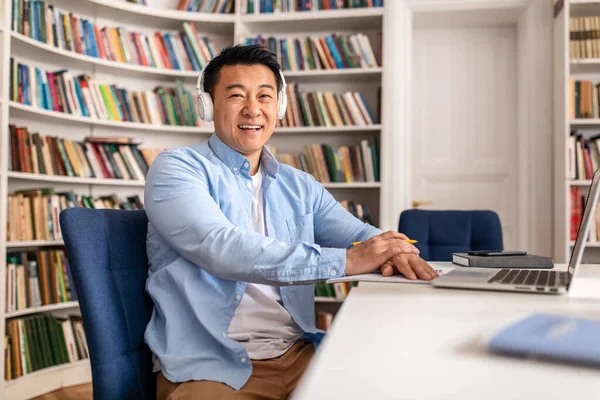 Happy Korean Male Teacher Near Laptop Posing Smiling To Camera Wearing Headphones Working And Teaching Online Sitting In Library Indoors. Modern Education And Technology Concept