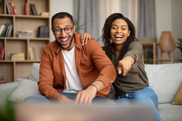 Joyful Black Couple Watching Funny Comedy Movie On TV Laughing And Pointing Finger Having Fun Sitting On Couch At Home On Weekend. Family Leisure And Entertainment Concept