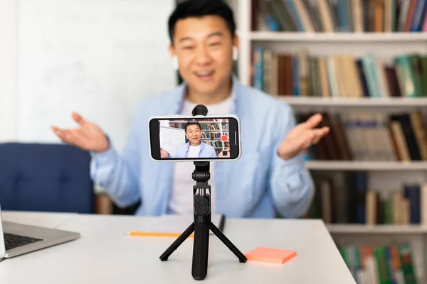 Korean Male Teacher Video Calling Using Cellphone On Tripod Communicating Online And Teaching Having Distance Class Sitting Indoor. Selective Focus On Mobile Phone. E-Teaching Concept