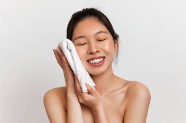 Yüz bakımı konsepti. Yüzü havluyla kuruyan, gözleri kapalı gülümseyen, beyaz stüdyo arka planında poz veren Koreli genç kadının portresi. Sabah güzellik rutini