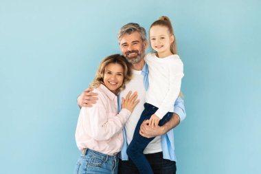 Mutlu Aile. Kızını kollarına alan ve karısını kucaklayan bir adam mavi stüdyoda yan yana duruyor. Küçük Çocuk Kameraya Gülümseyen Ebeveynler