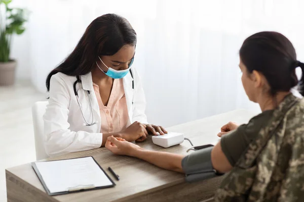 Black Doctor Lady In Protective Mask Measuring Blood Pressure For Military Woman, African American Medical Worker Checking Health Condition Of Female Soldier In Camouflage Uniform, Closeup Shot