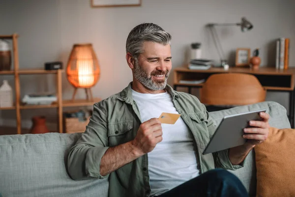 Smiling mature european man with beard with credit card looks at tablet and pay in living room interior. Shopping online, delivery and money, finance and technology at home during covid-19 quarantine