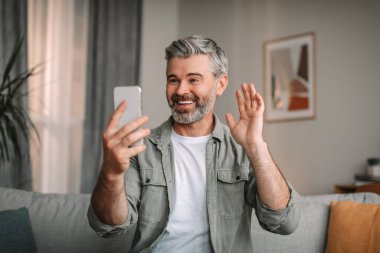 Oturma odasının içindeki akıllı telefon web kamerasına elini sallayan sakallı, gülümseyen yaşlı Avrupalı bir adam. Video çağrısı, uzaktan iletişim, yeni uygulama ve teknoloji covid-19 karantinasında.