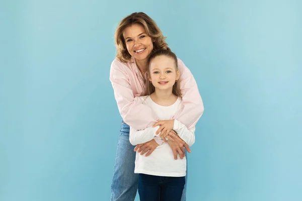 Joyful Mom Embracing Little Daughter Standing In Studio Over Blue Background. Mother Hugging Her Little Kid Smiling To Camera Posing Together. Family And Motherhood