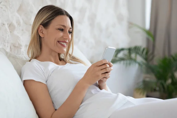 快乐的千年金发碧眼的女士坐在家里的床上 在智能手机上享受最新的购物手机软件 在网上买衣服和鞋子 复印空间和侧视图 — 图库照片