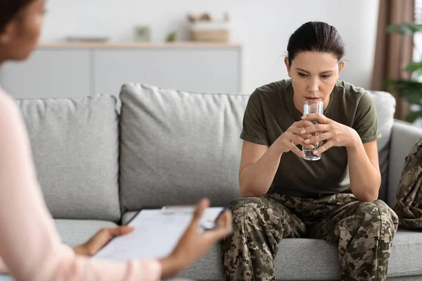 Portrait of stressed military woman at therapy session with black female psychologist, depressed soldier lady sitting on couch and holding glass with water, suffering mental problems or ptsd