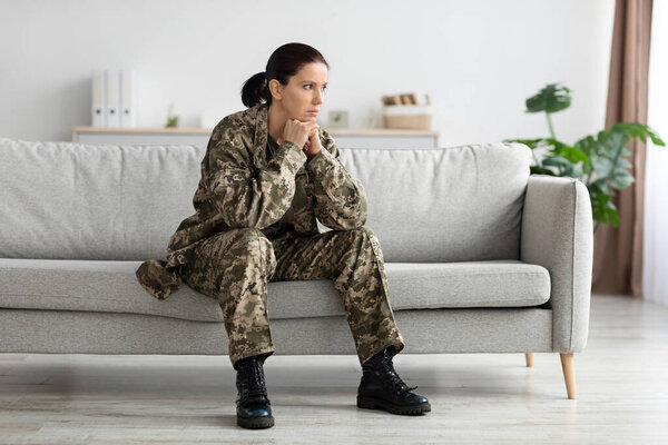 Депрессия после армии. Заботливая воительница в форме сидит на диване в помещении и смотрит в сторону, вдумчивая расстроенная военная женщина опираясь подбородок на руки, страдая психическими проблемами, копировать пространство