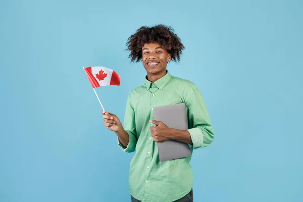 Modern education and student exchange. Happy black guy with laptop holding small flag of Canada and smiling at camera, standing over blue background, studio shot, copy space