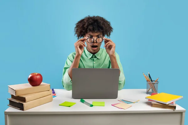 Omg. Portrait of shocked black guy in eyeglasses sitting at desk using laptop and looking at computer screen over blue studio background. Teenager reading shocking news
