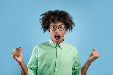 Heyecanlı siyahi adam başarıya seviniyor, yumruklarını sallıyor, mavi stüdyo arka planında heyecanla bağırıyor. Siyah erkek zaferi kutluyor, zaferi kutluyor, başarıyı kutluyor ve zaferi kutluyor.