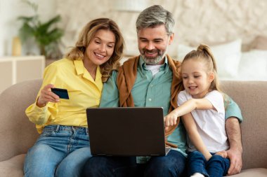 Mutlu Aile Alışverişi İnternette, ödeme için dizüstü bilgisayar ve kredi kartı kullanarak hafta sonu evdeki koltukta oturan bilgisayarı işaret eden çocuk. Ticaret Konsepti