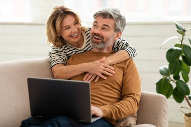 Neşeli orta yaşlı çift birlikte dizüstü bilgisayar kullanıyor internete bakıyor ve evdeki koltukta oturup film izliyor. Kadın bilgisayarda çalışırken kocasına sarılıyor. Aile ve Teknoloji
