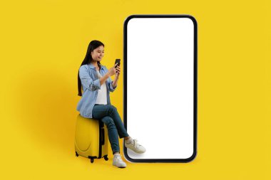 İyi ki genç Çinli bir kadın boş ekranlı kocaman bir telefonun yanında bavulun üzerine oturmuş sarı arka planda, stüdyoda izole edilmiş akıllı telefonuyla daktilo yazıyor. Tatilde seyahat, rezervasyon, online sohbet, blog, uygulama ve reklam