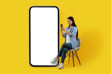 Gülümseyen bin yıllık Japon bayan, sarı arka planda izole edilmiş akıllı telefonuyla büyük ekranlı telefonun yanındaki sandalyeye oturuyor. Modern teknoloji, sosyal ağlarda online sohbet, blog, yeni uygulama ve reklam