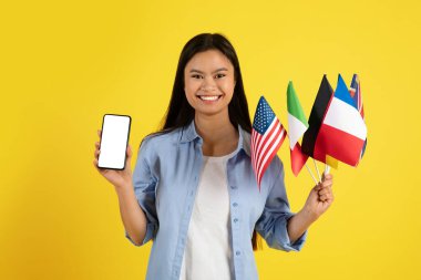 Pek çok farklı bayraklı neşeli genç Japon kız öğrenci sarı arka planda ve stüdyoda izole edilmiş boş ekranlı bir telefon gösteriyor. Öğrenim ve eğitim başvurusu, reklam, teklif, modern teknoloji