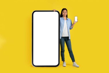 Yeni uygulama, dijital reklam ve teklif. Japon genç bayanın akıllı telefonu göstermesine sevindim. Sarı arka planda, stüdyoda, boş ekranı olan büyük bir telefonun yanında duruyor. Sosyal ağlarda, bloglarda, web sitelerinde çevrimiçi sohbet