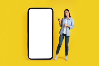 Sosyal ağlar ve sohbet. Neşeli mutlu Asyalı genç bayan büyük bir telefonu işaret ediyor. İlan için boş ekranı var, sarı arka planda, stüdyoda izole edilmiş. Yeni blog, uygulama, teklif ve modern teknoloji