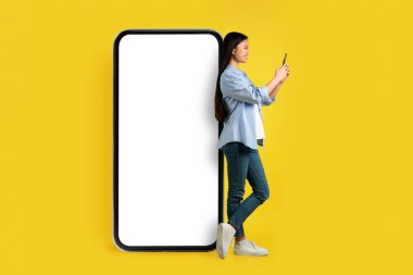 Akıllı telefonlu gülümseyen Asyalı genç bir kadın sarı arka planda, stüdyoda izole edilmiş boş ekranlı dev bir telefonun yanında duruyor. Sosyal ağlarda çevrimiçi sohbet, blog, uygulama, reklam, teklif ve modern teknoloji