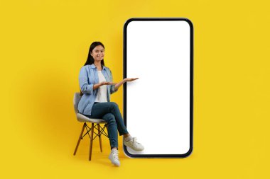 Neşeli genç Asyalı kadın sandalyeye oturur, büyük telefona el kaldırır, sarı arka planda boş ekran, stüdyo. Sunum, sosyal ağlarda online sohbet, blog, uygulama, reklam, teklif ve modern aygıt