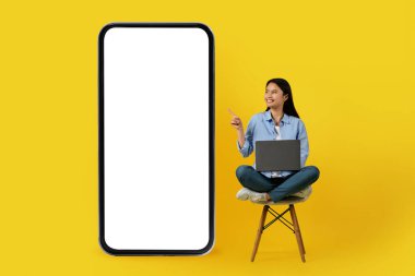 Genç ve mutlu Japon bayan dizüstü bilgisayarla sandalyede oturur ve sarı arka planda, stüdyoda izole edilmiş boş ekranlı akıllı telefonu işaret eder. Cihaz üzerinde yeni uygulamalarla sohbet et, online çalış, teklif et ve reklamını yap