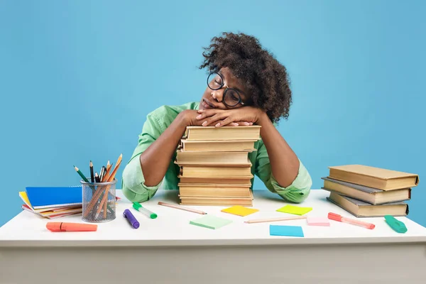 Exhausted black male student sleeping on desk on stacks of books, being tired while preparing for exam on blue studio background. African american teenager feeling overworked while studying