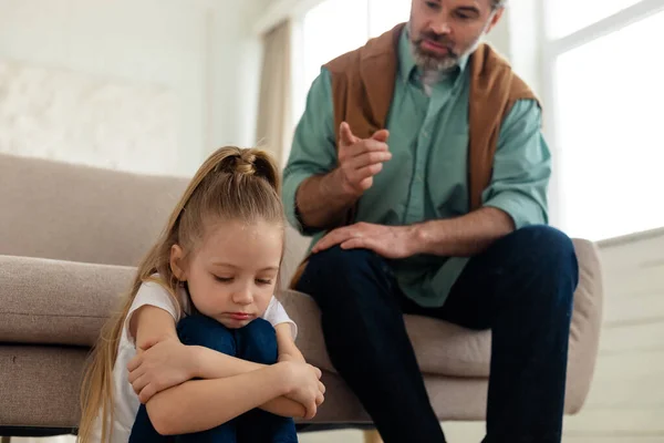 严格的中年爸爸闷闷不乐的小女儿而孩子抱着自己坐在地板上在家里 不幸的童年和父母问题概念 选择性焦点 — 图库照片