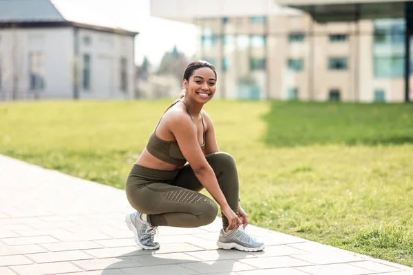 トレーニング前にスニーカーでレースを結ぶ楽しい若いアフリカ系アメリカ人女性 都会の公園でジョギング フリースペース アウトドアランニングのための陽気な黒人女性 アクティブなライフスタイルをリード — ストック写真