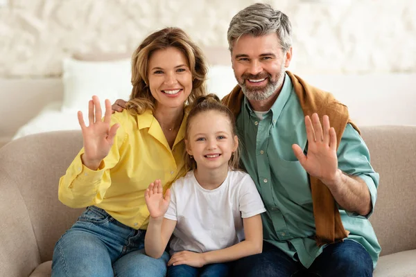 Cheerful Middle Aged Parents And Little Daughter Waving Hands Smiling To Camera Sitting On Sofa At Home. Family Of Three Gesturing Hi Greeting Indoor. Hello Concept