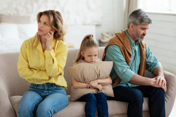 Unhappy Middle Aged Parents And Little Daughter Sitting Back To Back Not Speaking After Quarrel At Home, Frustrated Kid Hugging Pillow. Bad Family Relationship, Crisis And Problems Concept