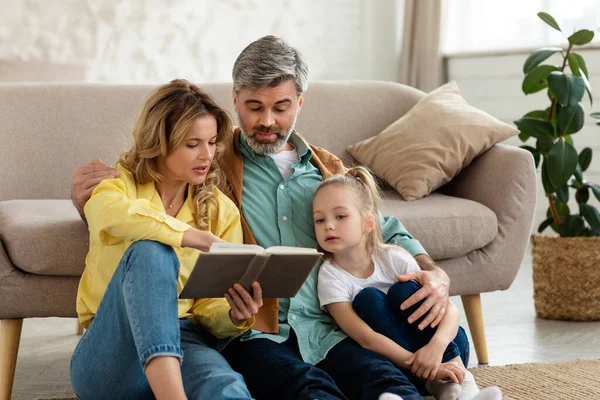 Family Reading Book Hugging Little Daughter Bonding Spending Time Together Sitting Near Couch At Home. Parents Read Fairytales With Kid. Weekend Leisure Concept