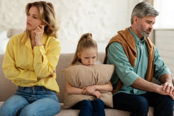 Unhappy Family Of Three Sitting Together Having Problems In Relationship At Home. Middle Aged Parents And Little Daughter Not Talking After Quarrel Indoors, Sad Kid Girl Embracing Pillow